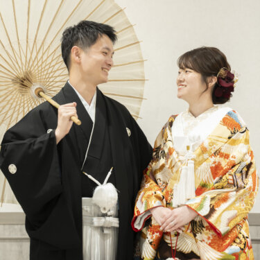 白い和傘を差して笑い合う黒紋付袴の新郎と、鶴柄のオレンジの色打掛の花嫁。横浜のスタジオで撮る和装前撮り