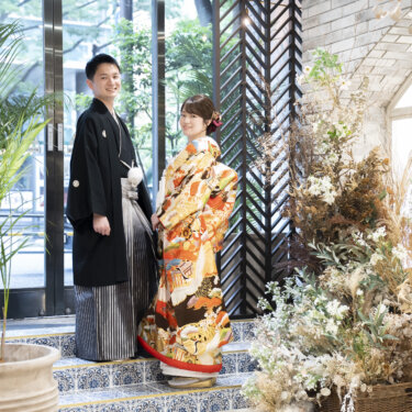 観葉植物とタイルの床のあるスタジオで並んで立つ黒紋付袴の新郎とオレンジの色打掛の花嫁。和装スタジオフォトの全身カット