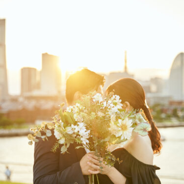 夕日の逆光の中、大きなブーケで顔を隠してキスする新郎新婦。遊び心のある顔隠しポーズのウェディングフォト
