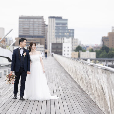 大さん橋のウッドデッキを手をつないで歩く新郎新婦。横浜の街並みを背景にした洋装ロケーションフォト
