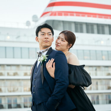 赤と青のラインの客船の前で、新郎の肩に手を添えて寄り添う黒いドレスの花嫁。港町らしい洋装前撮りのポーズ