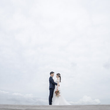 広い空を背にウッドデッキに小さく立つ新郎新婦を引きで撮った一枚。海と橋を遠くに望む洋装ロケーションフォト