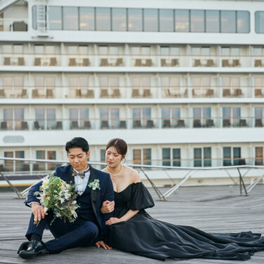 大型客船の前のウッドデッキに寄り添って座る新郎新婦。トレーンを広げた黒いカラードレスと大きなブーケが映える洋装前撮り