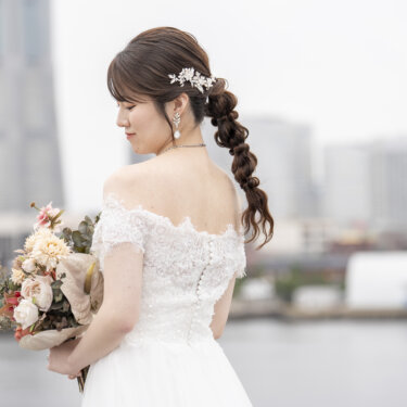 みなとみらいのビル群を背にブーケを抱えてうつむく花嫁の横顔。編み込みヘアと白い髪飾りが映えるオフショルダードレスの前撮りポートレート