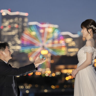 虹色に光る観覧車を背に、新郎が花嫁の手にそっと触れようとする一枚。みなとみらいの夜景を活かしたフォトウェディングの構図