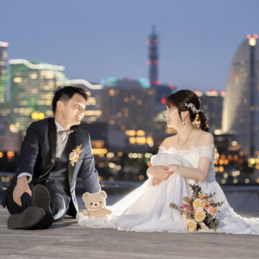 大さん橋のウッドデッキに座り、テディベアを間に置いて見つめ合う新郎新婦。夕暮れの横浜の夜景を背景にしたロケーション前撮り
