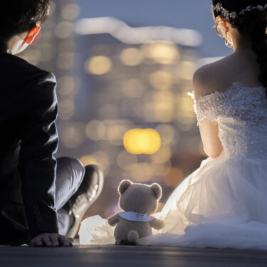 夜景のボケを背に並んで座る新郎新婦の後ろ姿と、間に置いた小さなテディベア。ぬいぐるみを使った夜の前撮りのアイテムカット
