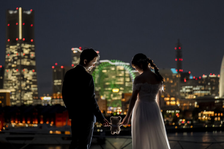 観覧車とランドマークタワーの夜景を背に、テディベアを手に手をつなぐ新郎新婦。横浜みなとみらいで撮る夜の前撮り