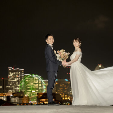 ランドマークタワーと観覧車の夜景を背に、ドレスの裾を広げて新郎と手をつなぐ花嫁。大さん橋の夜の前撮り