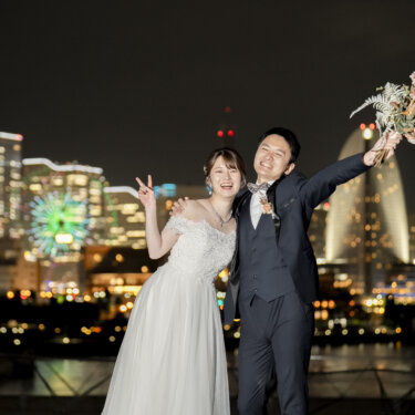 夜景を背にブーケを高く掲げて笑う新郎とピースをする花嫁。夜の前撮りで撮る元気なポーズ