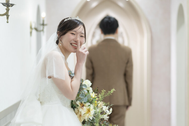 白いアーチの廊下で人差し指を口元に当ててほほえむ花嫁と、後ろ姿の新郎。ベールとティアラのウェディングドレスで撮る横浜のスタジオフォト