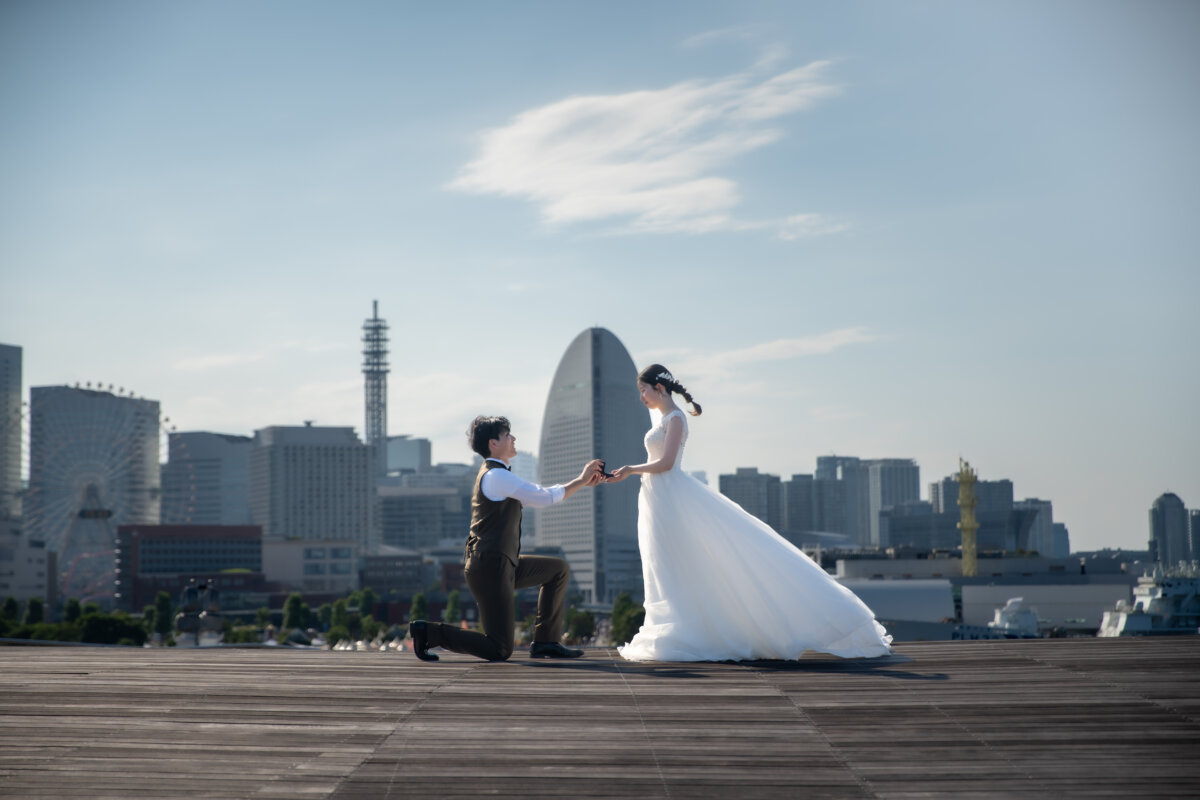 コスモクロック21とランドマークタワーを背に、片膝をついて花嫁の両手を取る新郎。海風にドレスがなびくプロポーズ再現の洋装前撮り