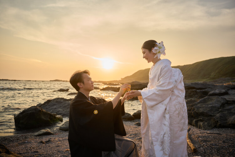 城ヶ島の岩場で夕日を背に、ひざまずいて白無垢の花嫁の手を取る紋付袴の新郎。サンセットを生かした海辺の和装前撮り