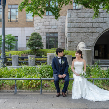 石造りの建物を背に歩道の柵に腰かけて見つめ合う新郎新婦。ロングトレーンのドレスが映える横浜の街ロケ前撮り