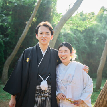 松の木立を背に肩を抱き寄せて笑う紋付袴の新郎と白無垢の花嫁。自然体の笑顔を残す和装フォトウェディング