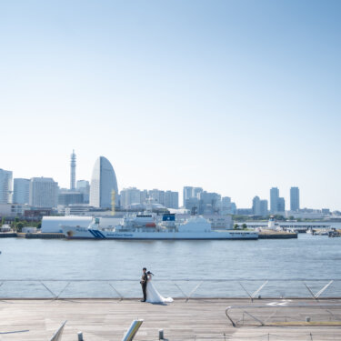 青空と港の広い景色の中に小さく佇む新郎新婦。大さん橋からみなとみらいを望む、引きの構図で撮るロケーションフォト