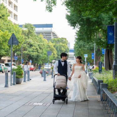 青い街路旗が並ぶ並木道を、ベビーカーを押して歩く新郎新婦を引きで写した一枚。家族で歩く横浜の街ロケーションフォト