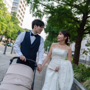 並木道でベビーカーを押しながら見つめ合う、ベスト姿の新郎とウェディングドレスの花嫁。子連れで楽しむ街歩きの前撮り