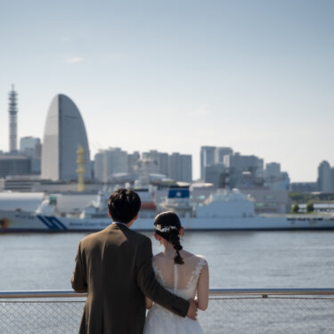 港の向こうに広がる横浜の街並みを並んで見つめる新郎新婦の後ろ姿。ヘアアレンジの髪飾りが光る洋装前撮りのワンシーン