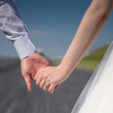 グレーのシャツの新郎と結婚指輪をはめた花嫁が指先をつなぐ手元アップ。ウェディングドレスのチュールが写り込む前撮りのディテール