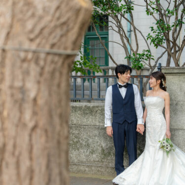 大きな木の幹の向こう、石塀の前で手をつないで並ぶ新郎とマーメイドドレスの花嫁。街歩きで撮る洋装ロケーションフォト