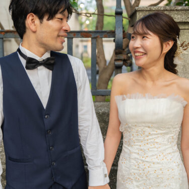 ベストに蝶ネクタイの新郎と手をつなぎ、見つめ合って笑うチュールのウェディングドレスの花嫁。街歩きで撮る洋装前撮り