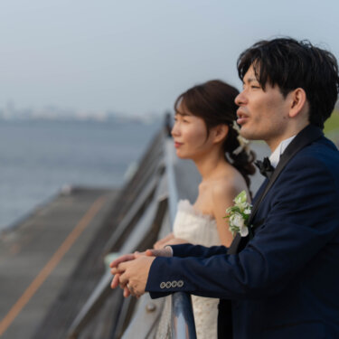 海を眺めるデッキの柵に並んでもたれ、言葉を交わす新郎新婦の横顔。曇り空の下でも雰囲気のある洋装前撮り