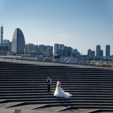 帆の形のホテルが建つみなとみらいの街並みを望む階段で、ブーケを持つ花嫁と並ぶ新郎。広い空が開放的な洋装前撮り