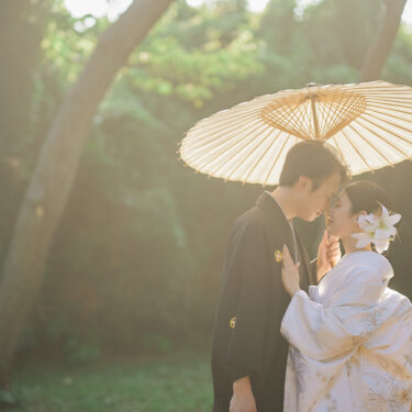 逆光の松林で和傘を差し、額を寄せ合う紋付袴の新郎と白無垢の花嫁。城ヶ島で撮るやわらかな光の和装前撮り