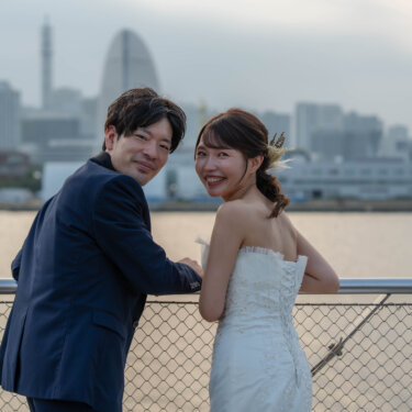 みなとみらいの街並みを背に振り返って微笑む新郎新婦。大さん橋の手すりにもたれて撮る洋装前撮りのナチュラルなカット
