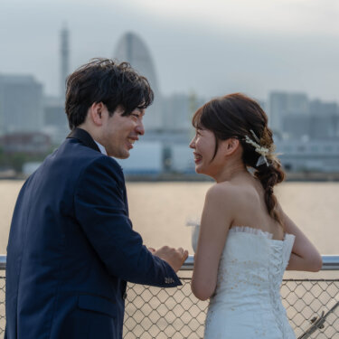 ランドマークタワーを望むデッキで笑い合うネイビーのタキシードの新郎とウェディングドレスの花嫁。みなとみらいの眺めが広がる前撮り