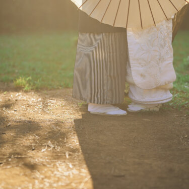 和傘の下に並ぶ紋付袴の新郎と白無垢の花嫁の足元。木漏れ日の地面に影が伸びる和装前撮りのディテールカット