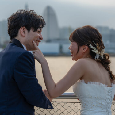 フェンス越しの海を背に、新郎の頬に手を添えて見つめ合う花嫁。マーメイドラインのドレスが大人っぽい洋装ロケーションフォト
