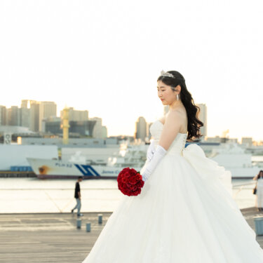 赤いバラのブーケと白いグローブを合わせたプリンセスラインのウェディングドレスの花嫁が、港を見つめる横顔の前撮り