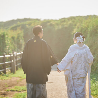 柵の続く城ヶ島の遊歩道を手をつないで歩く紋付袴の新郎と白無垢の花嫁の後ろ姿。海辺の和装ロケーションフォト