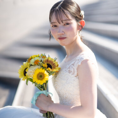 ひまわりのブーケを抱えて座り、こちらを見つめるウェディングドレスの花嫁。夏のブーケが映える花嫁ソロの前撮りポーズ