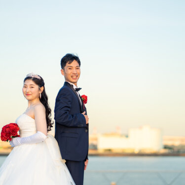 港を背景に背中合わせで立ち、赤いバラのブーケを持って笑う新郎新婦の定番ポーズの結婚写真