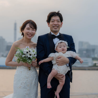 横浜・大さん橋でみなとみらいのビル群を背に、赤ちゃんを抱くネイビーのタキシードの新郎とブーケを持つ花嫁の家族写真。子連れの洋装前撮り