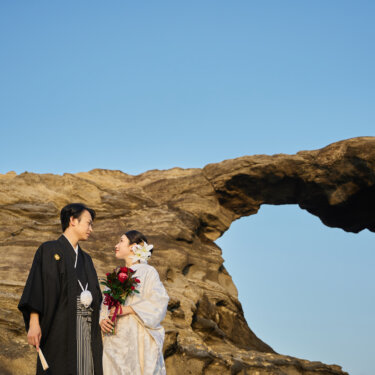 城ヶ島の馬の背洞門の岩のアーチを背に、赤いバラのブーケを持つ白無垢の花嫁と紋付袴の新郎。絶景の和装ロケーションフォト