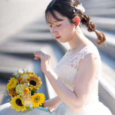 編み込みのヘアアレンジに小花の髪飾りをつけ、ひまわりのブーケを手に目を伏せる花嫁。逆光のやわらかな光の中で撮るウェディングフォト