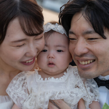 赤ちゃんを真ん中に両側からほおを寄せる新郎新婦の親子三人のアップ。笑顔あふれる家族写真のフォトウェディング