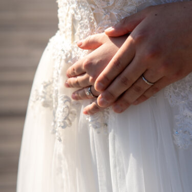 結婚指輪をはめた新郎新婦の手が、レース刺繍のウェディングドレスの前でそっと重なる手元アップの前撮り