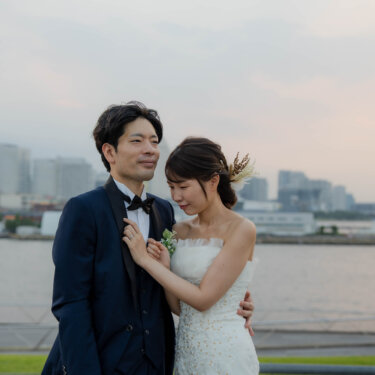 ビル群と海を背に寄り添い、新郎の胸元に手を添えて微笑むマーメイドドレスの花嫁。横浜の海辺で撮る洋装ロケーションフォト