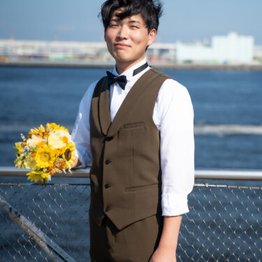 ひまわりのブーケを片手に、海を背にして手すりの前に立つブラウンのベスト姿の新郎。夏らしい爽やかな新郎ソロの前撮りカット