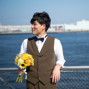 ひまわりのブーケを持ち、海を背に手すりにもたれて微笑む蝶ネクタイの新郎。港の青空が爽やかな夏のフォトウェディング