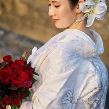 夕日に照らされた岩場で、ユリの髪飾りを付けて赤いバラのブーケを抱く白無垢の花嫁の横顔。和装前撮りの花嫁ソロカット