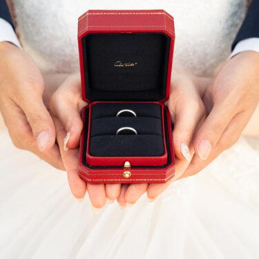 ドレスの上で新郎新婦が両手を重ね、赤いリングボックスの結婚指輪を見せる手元のウェディングフォト