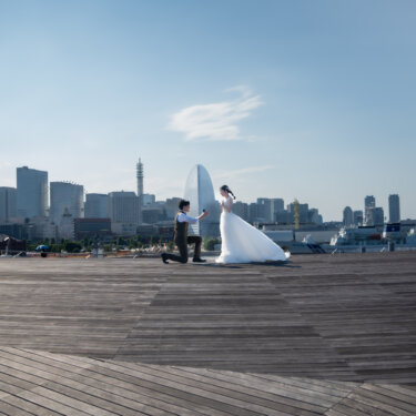 横浜の高層ビルを望む広いデッキで、ひざまずいて花嫁に手を差し出す新郎。青空の下のプロポーズシーンを引きで撮ったウェディングフォト