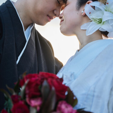 夕方の光の中、おでこを寄せて目を閉じる紋付袴の新郎と赤いブーケを持つ白無垢の花嫁。おでこ合わせポーズの和装前撮り