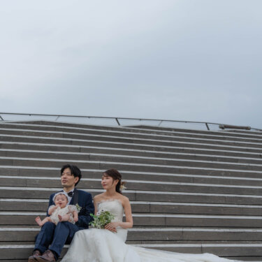 空を大きく入れて階段の下に小さく座る親子三人を写した一枚。余白を生かした家族フォトウェディングの構図
